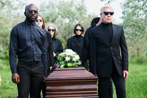 Group of grieving people in mourning attire carrying wooden coffin with bunch of fresh white chrysanthemums on top of closed lid at funeral